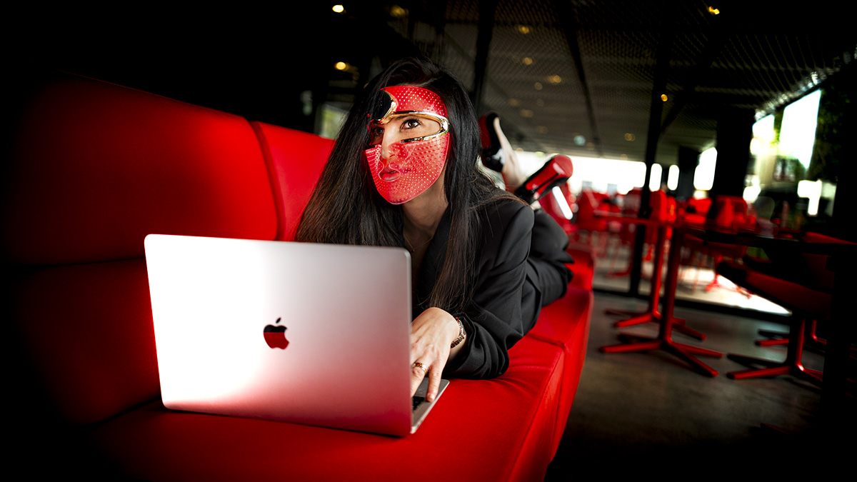 A dark-haired woman, lying on her stomach on a blood-red couch in an elegant, modern glass and metal coffee shop with red tables, working on a laptop and wearing the LED face mask